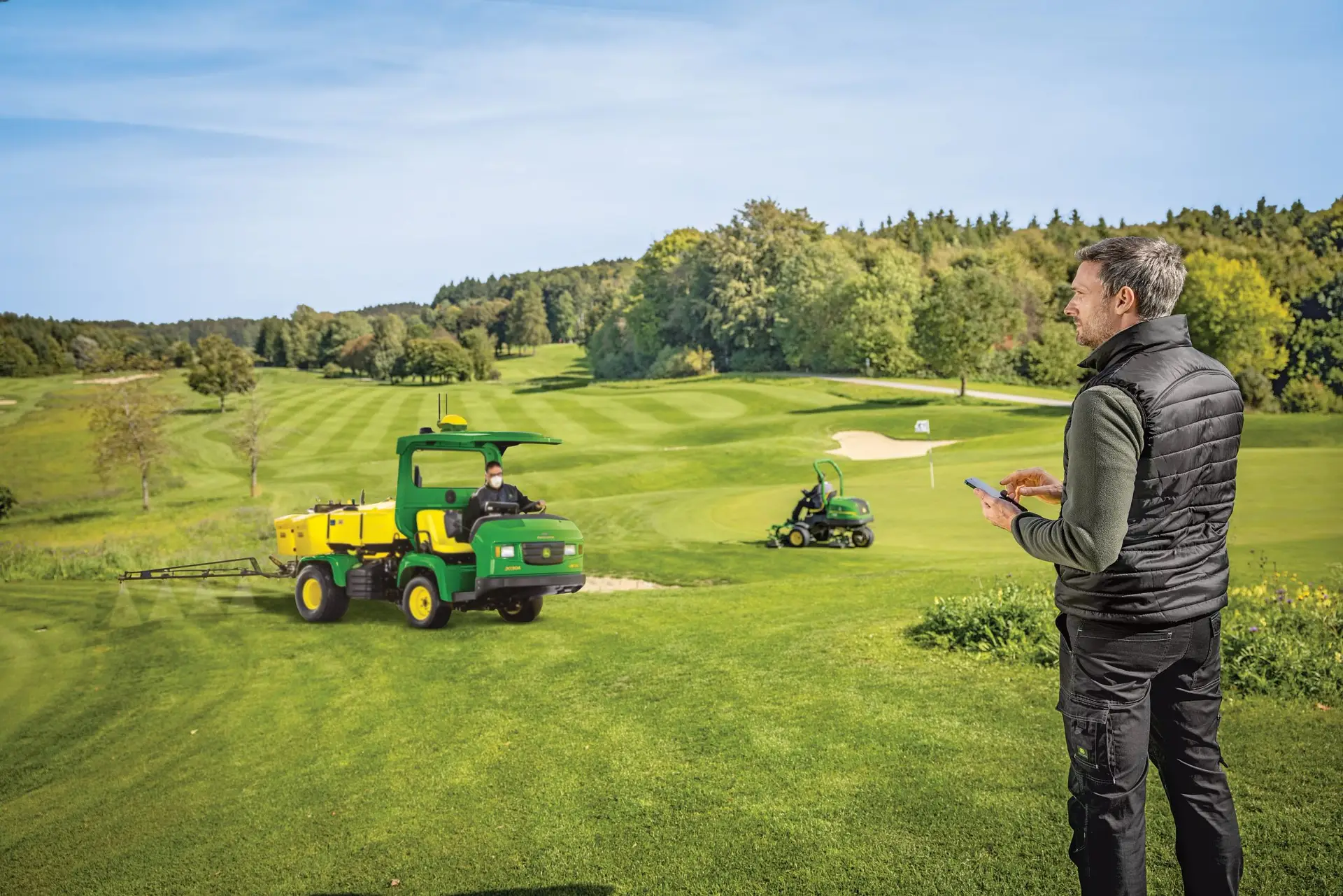 Mann mit Handy vor Golfplatz, John Deere Pflegemaschinen auf der Anlage